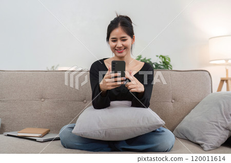 Cute Young Woman Enjoying a Relaxing Moment Playing on Her Smartphone While Sitting on a Cozy Sofa at Home 120011614