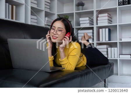 Young Woman Relaxing Happily in Modern Living Room with Laptop and Headphones, Enjoying Leisure Time on Comfortable Black Sofa 120011640