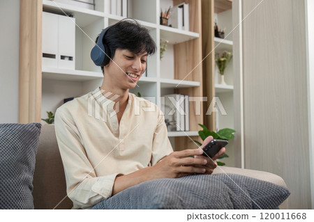 Young Man Relaxing in Modern Room While Playing on Phone and Wearing Headphones Young Man Relaxing in Modern Room While Playing on Phone and Wearing Headphones 120011668