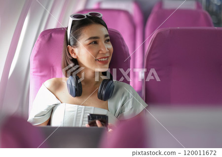 Smiling Woman Traveling by Plane with Headphones and Smartphone, Seated in Modern Airplane Cabin with Pink Seats Smiling Woman Traveling by Plane with Headphones and Smartphone, Seated in Modern Airplane Cabin with Pink Seats 120011672