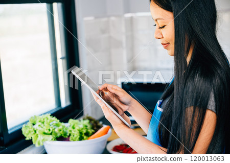 In a sunny kitchen a cheerful Asian woman joyfully prepares a meal using a tablet for cooking. Illustrating the harmonious blend of technology culinary artistry and domestic bliss. 120013063