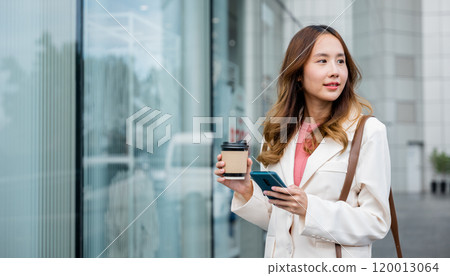 Young businesswoman on her way to the office, multitasking with a cup of coffee and a smartphone, balancing work and personal life. 120013064