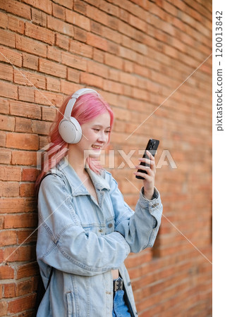 A girl with pink hair is standing next to a brick wall, holding a cell phone 120013842