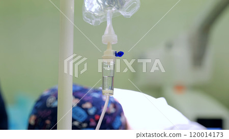 Patient in surgical cap lies in hospital bed with iv drip in focus, background blurred Patient in surgical cap lies in hospital bed with iv drip in focus, background blurred 120014173