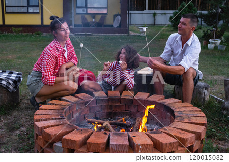 Family mom, dad, daughter roast marshmallows on skewers on a campfire in the hearth in the courtyard of the house by the fire pit on a cozy summer evening. 120015082
