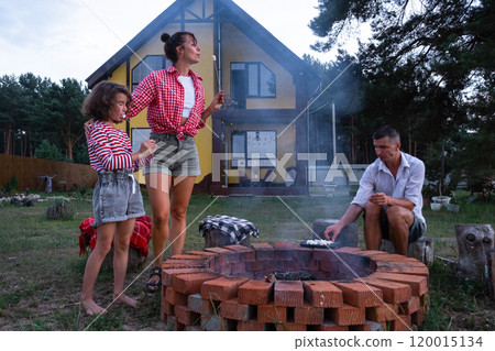 Family mom, dad, daughter roast marshmallows on skewers on a campfire in the hearth in the courtyard of the house by the fire pit on a cozy summer evening. Family mom, dad, daughter roast marshmallows on skewers on a campfire in the hearth in the courtyard of the house by the fire pit on a cozy summer evening. 120015134