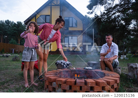 Family mom, dad, daughter roast marshmallows on skewers on a campfire in the hearth in the courtyard of the house by the fire pit on a cozy summer evening. 120015136