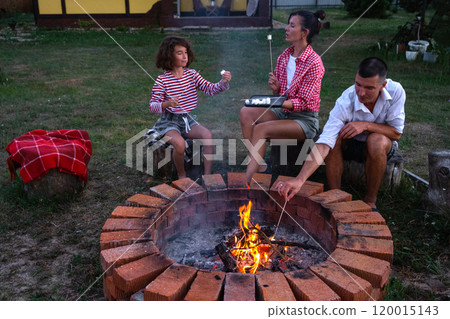 Family mom, dad, daughter roast marshmallows on skewers on a campfire in the hearth in the courtyard of the house by the fire pit on a cozy summer evening. 120015143