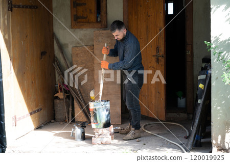 A man stirs liquid bitumen in a bucket with a wooden stick. 120019925