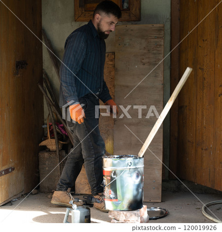 A man stirs liquid bitumen in a bucket with a wooden stick. 120019926