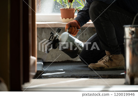 Waterproofing the floor in the house, a man heats a bitumen roll with a kerosene lamp. Waterproofing the floor in the house, a man heats a bitumen roll with a kerosene lamp. 120019946