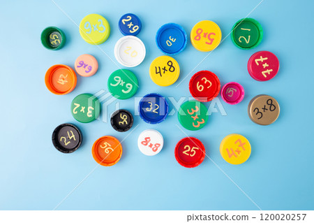 Colorful bottle caps with multiplication problems on blue background, flat lay math learning, engaging classroom activities, multiplication table practice, fun math activity for kids, home learning, 120020257
