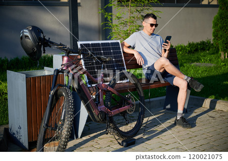 Man cyclist using solar panel for charging electric mountain bike outdoors. Guy in sunglasses sits on bench, holding phone, casually dressed in t-shirt, shorts, enjoying sunny outdoor setting. 120021075