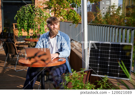 Man works on laptop and smartphone at outdoor table, with glass of iced coffee. Solar panel nearby, emphasizing sustainable, modern eco-friendly workspace that combines technology and sustainability. Man works on laptop and smartphone at outdoor table, with glass of iced coffee. Solar panel nearby, emphasizing sustainable, modern eco-friendly workspace that combines technology and sustainability. 120021089