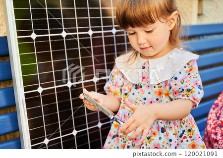 Young girl holds smartphone connected to solar panel for charging. Integration of sustainable renewable energy into daily life, importance of teaching children about eco-friendly technology. Young girl holds smartphone connected to solar panel for charging. Integration of sustainable renewable energy into daily life, importance of teaching children about eco-friendly technology. 120021091