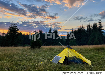 Yellow tent pitched in grassy field with solar panel beside it, capturing last light of setting sun. Backdrop of silhouetted trees and colorful sky emphasizes eco-friendly camping in nature. Yellow tent pitched in grassy field with solar panel beside it, capturing last light of setting sun. Backdrop of silhouetted trees and colorful sky emphasizes eco-friendly camping in nature. 120021094