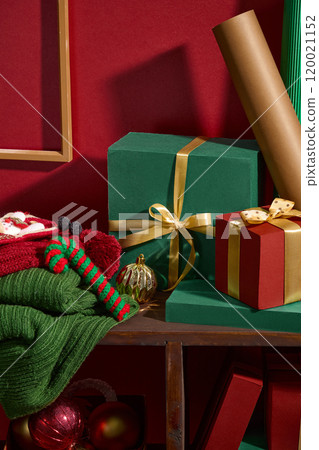 The image is a close-up shot of a still-life cozy Christmas featuring gifts, a stack of knit sweaters, a candy cane, and several Christmas ornaments placed on the wooden shelf in the red background. 120021152