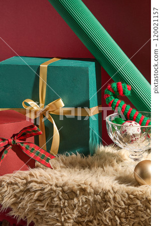 Christmas photo template with empty space in the middle to display product on a fur rug, next to it are glass cups containing Noel balls, a wool candy cane, and gift boxes, behind is a red background. Christmas photo template with empty space in the middle to display product on a fur rug, next to it are glass cups containing Noel balls, a wool candy cane, and gift boxes, behind is a red background. 120021157