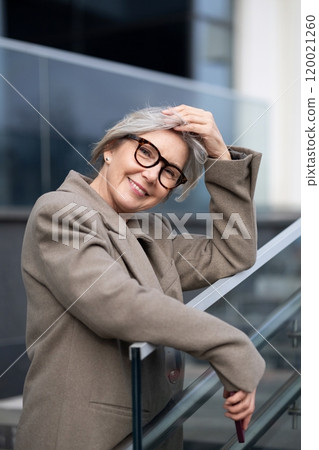 Middle-aged businesswoman smiles while leaning against a glass railing at an outdoor corporate Middle-aged businesswoman smiles while leaning against a glass railing at an outdoor corporate 120021260
