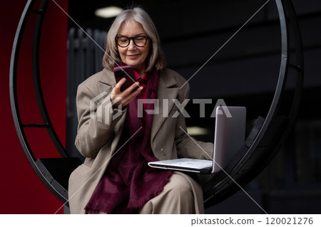 Senior businesswoman working outside on a laptop while checking her smartphone in a modern urban 120021276