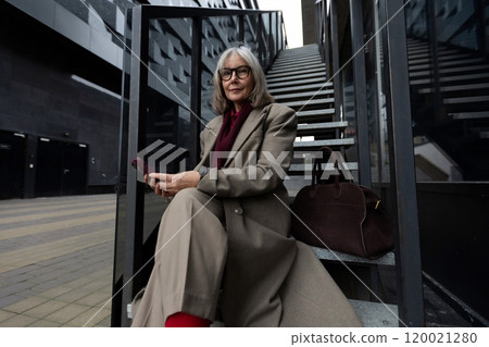 A middle-aged businesswoman seated on stairs, checking her phone outside a modern building on a A middle-aged businesswoman seated on stairs, checking her phone outside a modern building on a 120021280