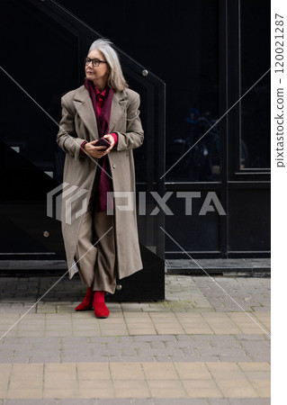 Stylish elderly woman with gray hair dressed in a long coat and red shoes stands gracefully against Stylish elderly woman with gray hair dressed in a long coat and red shoes stands gracefully against 120021287
