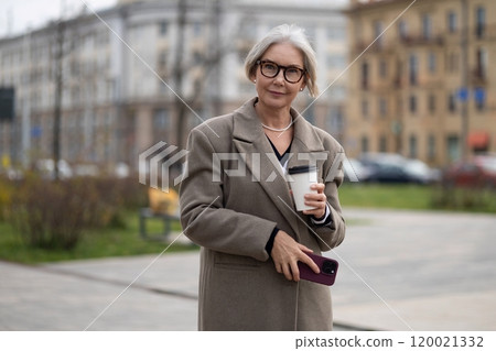 A senior businesswoman stands on a city sidewalk with a coffee cup and smartphone during a spring A senior businesswoman stands on a city sidewalk with a coffee cup and smartphone during a spring 120021332