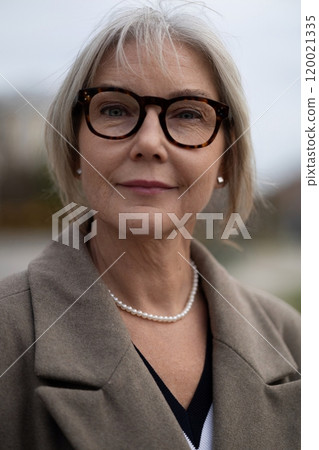 Elderly businesswoman in a stylish coat and glasses poses confidently outdoors on a cloudy day 120021335