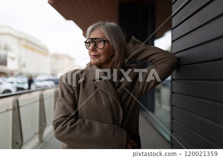 An older woman in a stylish coat leans against a modern building while enjoying a cloudy day in the An older woman in a stylish coat leans against a modern building while enjoying a cloudy day in the 120021359