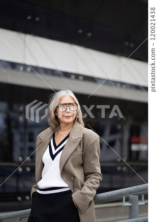 Elderly businesswoman standing confidently outside her office in professional attire during a sunny Elderly businesswoman standing confidently outside her office in professional attire during a sunny 120021438