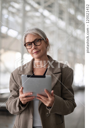 Elderly businesswoman standing confidently outside an office building, engaging with a tablet during 120021452