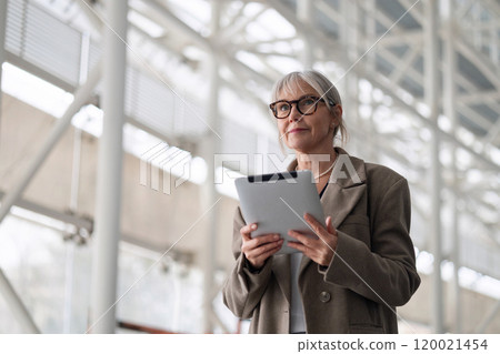 Elderly businesswoman standing confidently outside her office while holding a tablet and preparing 120021454