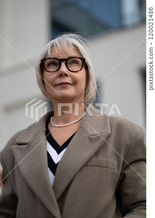 Stylish elderly woman with glasses and a chic coat standing confidently outdoors in a modern urban 120021486