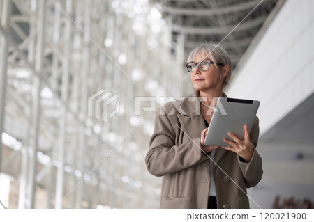 Stylish elderly woman with glasses using a tablet in a modern, well-lit architectural space during 120021900