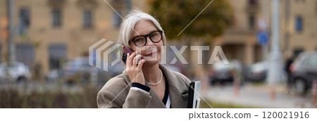 Elderly businesswoman on a phone call outside her office during a sunny afternoon in an urban Elderly businesswoman on a phone call outside her office during a sunny afternoon in an urban 120021916