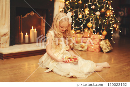 Happy little girl child with festive gift box sitting near Christmas tree and fireplace at home Happy little girl child with festive gift box sitting near Christmas tree and fireplace at home 120022162