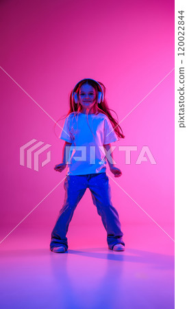 Dynamic photo of little charming girl dancing with joy while listening music in headphones in neon light against vivid gradient background. Dynamic photo of little charming girl dancing with joy while listening music in headphones in neon light against vivid gradient background. 120022844