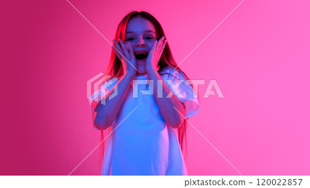 Girl with surprised expression holding hands on cheeks and looking excited in neon light against vivid gradient background. 120022857