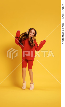 Girl in red costume, joyfully jumping with arms up, wearing headphones and standing against yellow studio background. 120022874