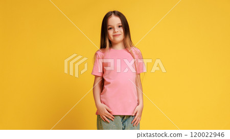 Stylish little girl, dressed casual attire, with smiling expression looking at camera, posing against yellow studio background. 120022946