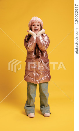 Happy little child, girl dressed winter clothes, smiling and holding hand near face against yellow studio background. Happy little child, girl dressed winter clothes, smiling and holding hand near face against yellow studio background. 120022957