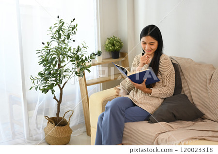 Beautiful young woman enjoys quiet time with a book in living room Beautiful young woman enjoys quiet time with a book in living room 120023318