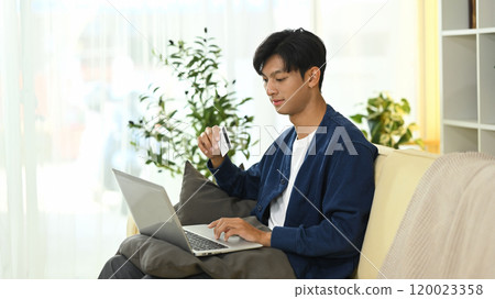 Handsome asian man holding credit card making purchase via laptop 120023358