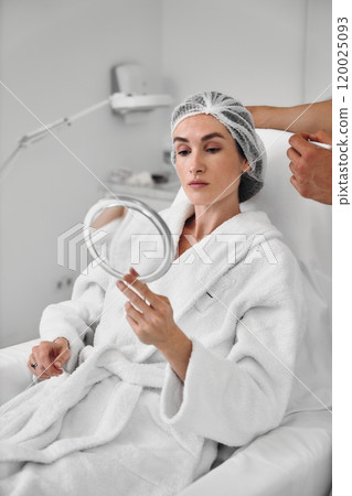 Woman in a spa robe examining her reflection in a mirror during treatment 120025093