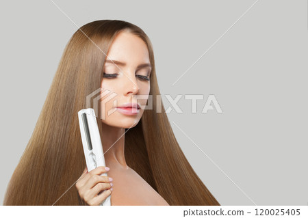 Stylish brunette woman holding straight hair iron and straightening her long smooth shiny hair  120025405