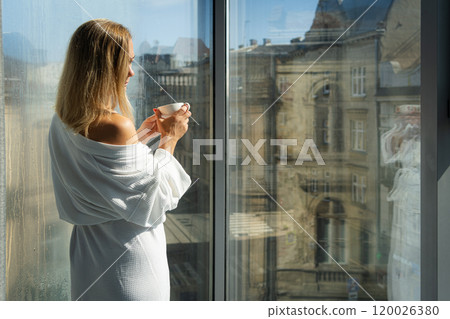 Woman in elegant robe drinking coffee in hotel room and standing near window Woman in elegant robe drinking coffee in hotel room and standing near window 120026380