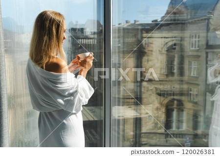 Woman in elegant robe drinking coffee in hotel room and standing near window Woman in elegant robe drinking coffee in hotel room and standing near window 120026381