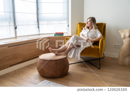 woman in a white dress sits on a chair in a hotel and looks out the window 120026389