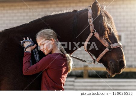 Young little teenager kid girl portrait hugging chestnut horse  at stable ranch yard. Horse riding school exercise. Cute little beginner blond girl near beautiful brown pet. Equine hobby therapy 120026533
