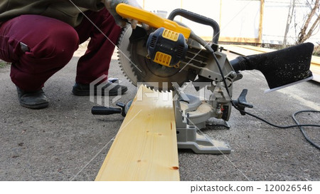 A worker wearing gloves saws a light-colored wooden board with a hand-held circular saw on the pavement. 120026546